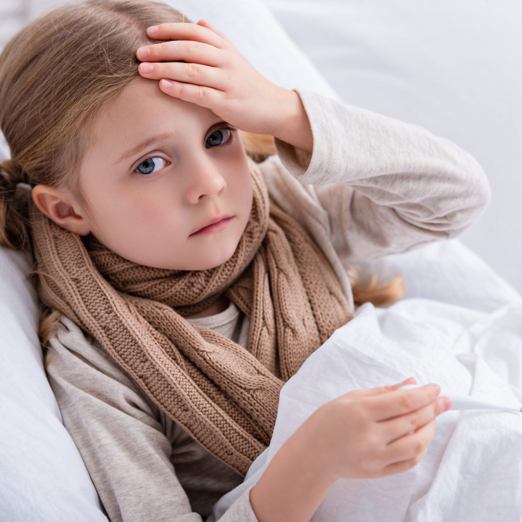 sick child with scarf over neck lying in bed, holding thermometer and touching forehead at home